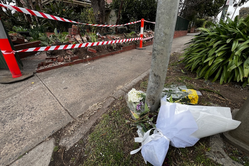 Bouquets of flowers lie on the pavement beside emergency services tape over a crushed brick fence.