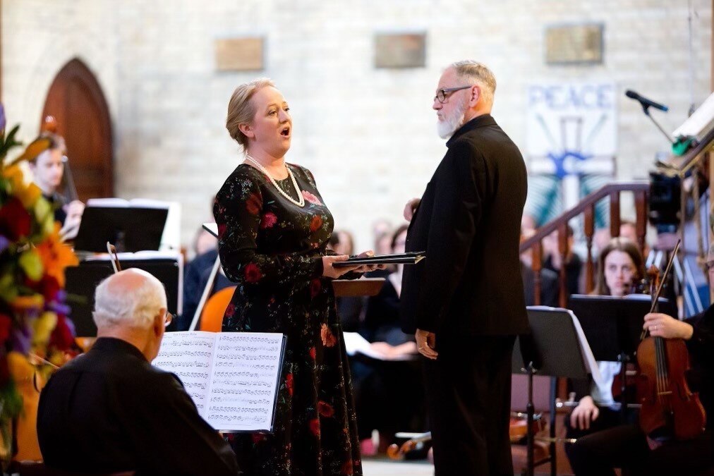 A female singer performs onstage, watched by an older man.