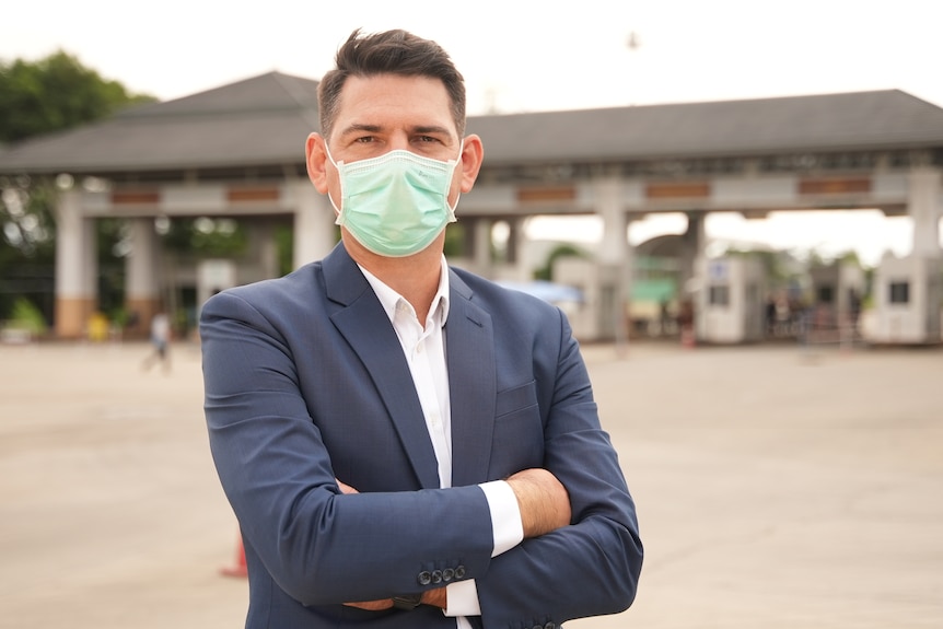 A man wearing a suit and disposable face mask stands with his arms crossed in front of a vehicle checkpoint