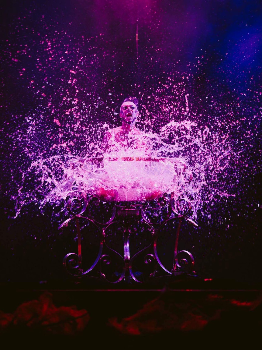 A man splashes in a bird bath on stage while lit by purple lights.