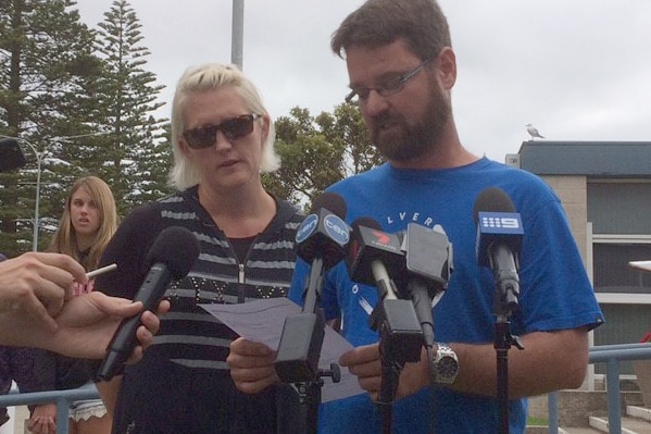 A mid shot of a man in a blue shirt reading a statement from paper with a woman alongside him and microphones in front.