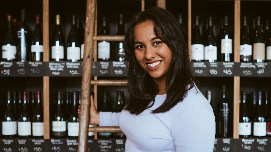 A woman stands on a ladder wearing a grey dress and looks to the camera smiling. Racks of wine are behind her.