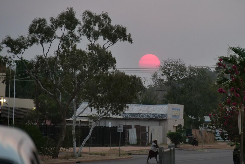 A pink sun setting through thick grey smoke, behind homes on a street in the town of Tennant Creek.
