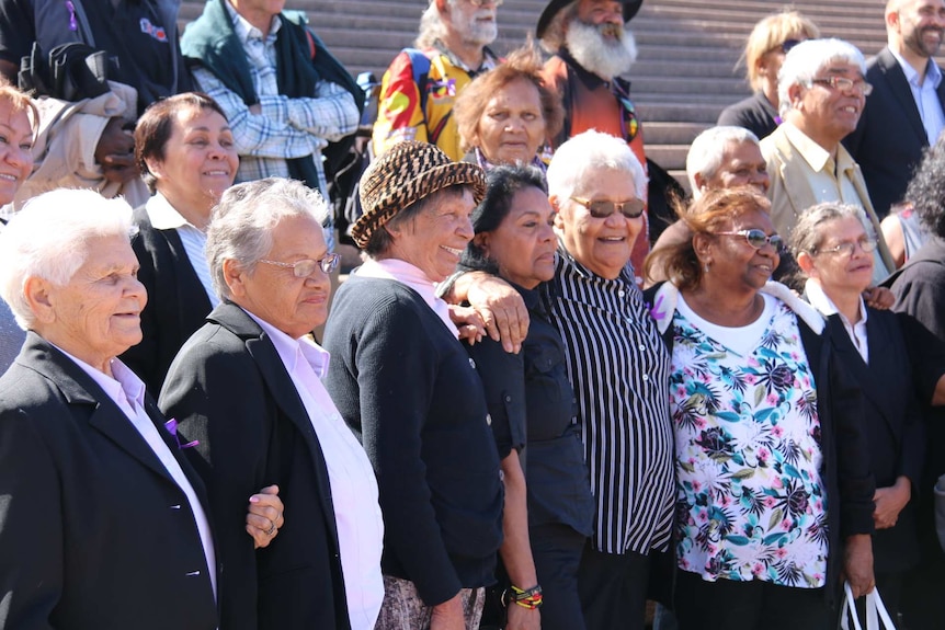 A group of more than a dozen Stolen Generations members are smiling on the steps outside the Sydney Opera House.