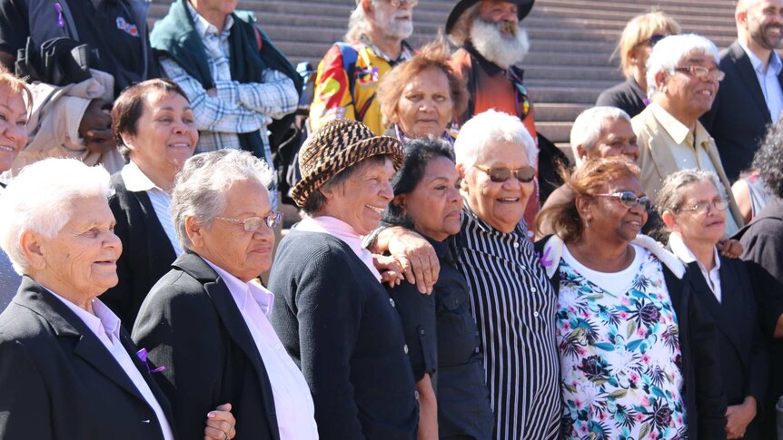 A group of more than a dozen Stolen Generations members are smiling on the steps outside the Sydney Opera House.