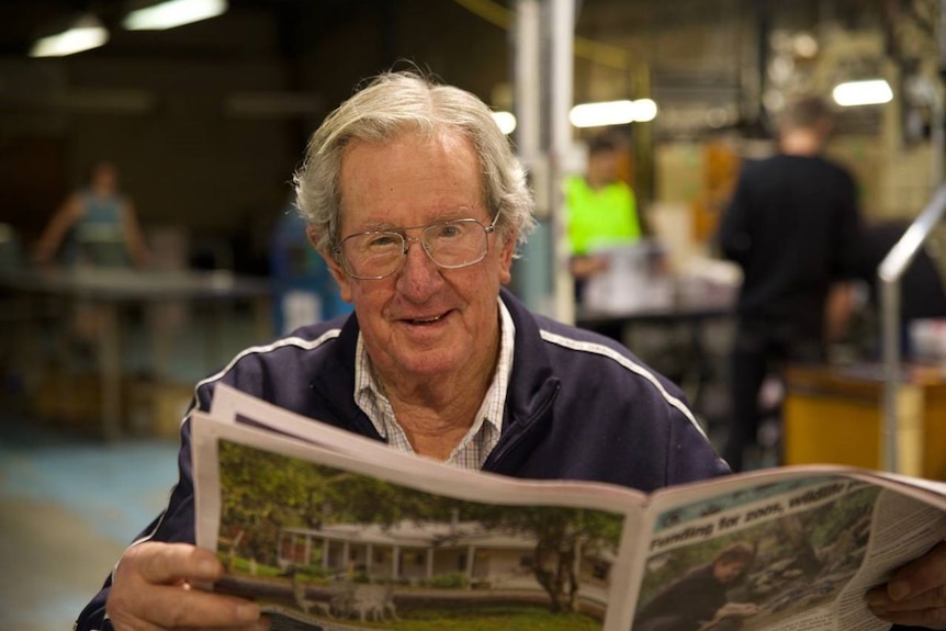 An elderly man in a blue jacket and glasses holds up a newspaper.