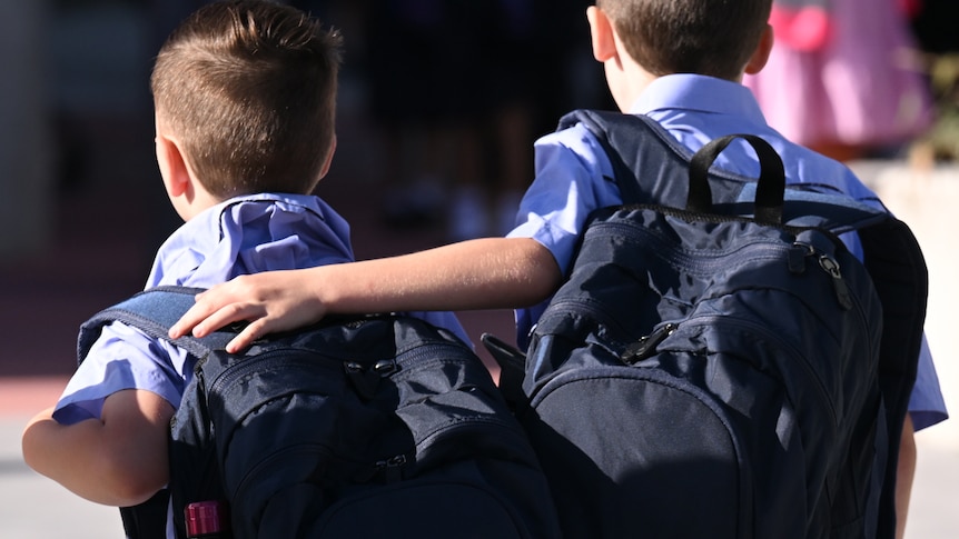 two young school boys walk to school wearing backpacks as one puts his arm around the others shoulder