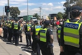 At least 20 police officers line the pavement as protestors stand in an intersection. Some protesters are holding signs.