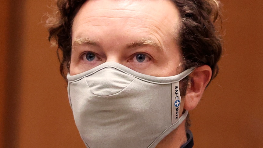 Danny Masterson wears a face mask looking straight ahead in front of an orange background.