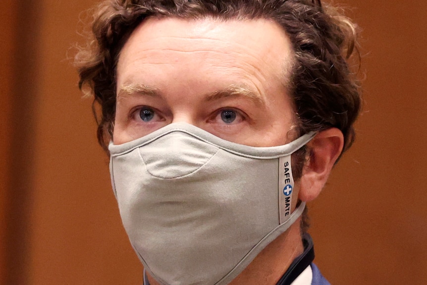 Danny Masterson wears a face mask looking straight ahead in front of an orange background.