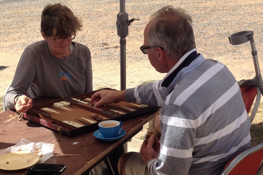 A man and a woman play backgammon under an umbrella, outside the Collector Community Hall.