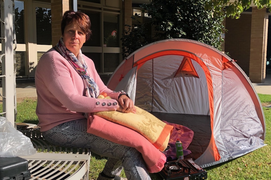 A woman sits in front of a tent, holding some blankets