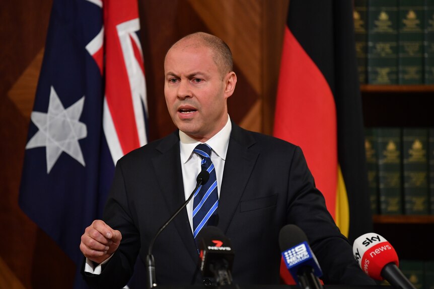 federal treasurer josh frydenberg speaks at a lectern covered in microphones in front of australian and aboriginal flags