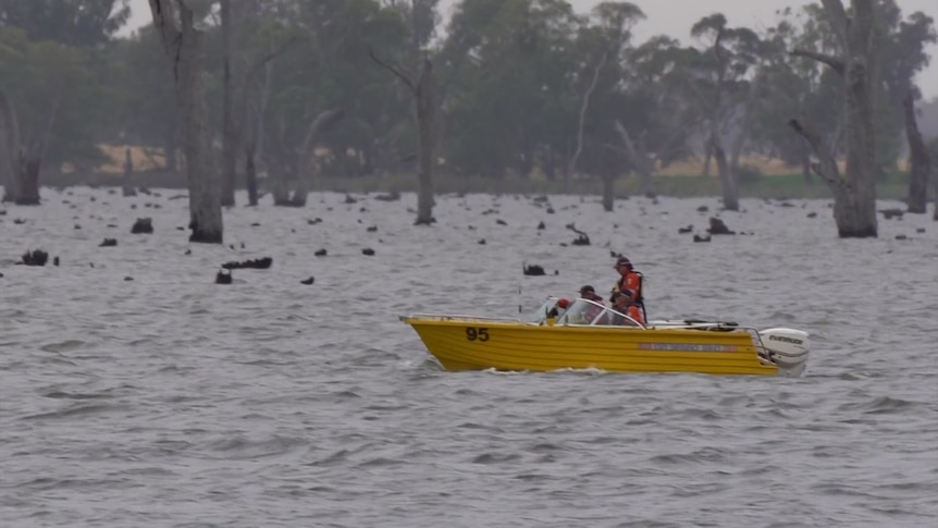 A yellow boat with two men in high visibility clothing on board travels on a grey lake.