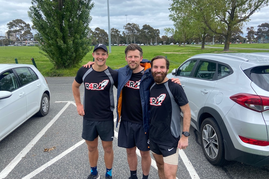 Three smiling men in matching T-shirts pose next to each other in a car park.