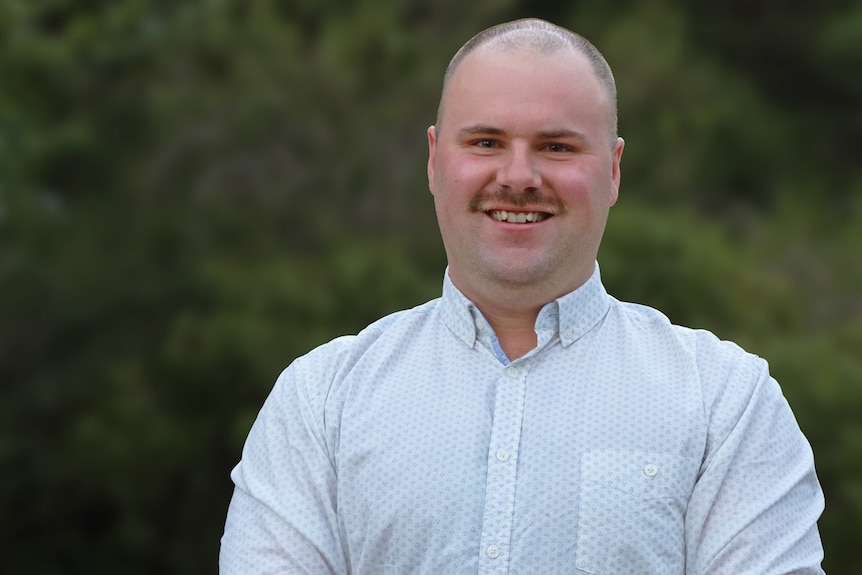 A smiling man with close-cropped hair and a moustache stands outside, smiling.