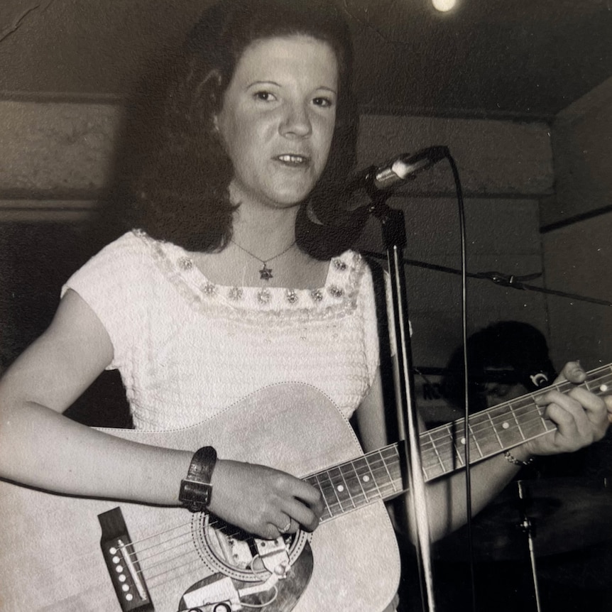 black and white historic photo of 70s styled teen girl at microphone holding guitar
