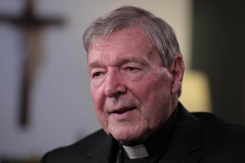 Cardinal George Pell sits in a chair wearing a clerical collar. A crucifix is seen in the background.