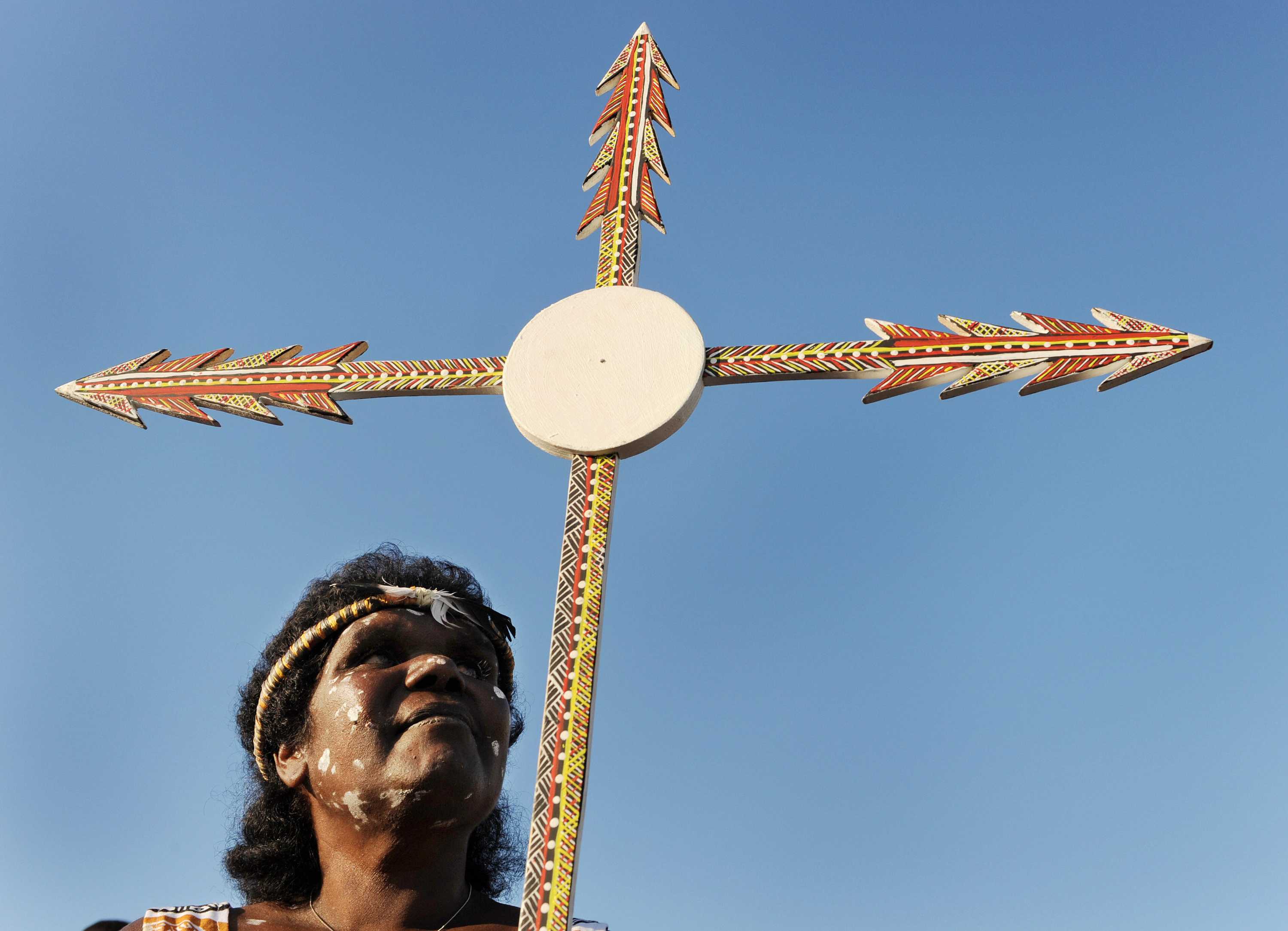 An Aboriginal woman holding a Christian cross made out of spears and painted in an Indigenous style.