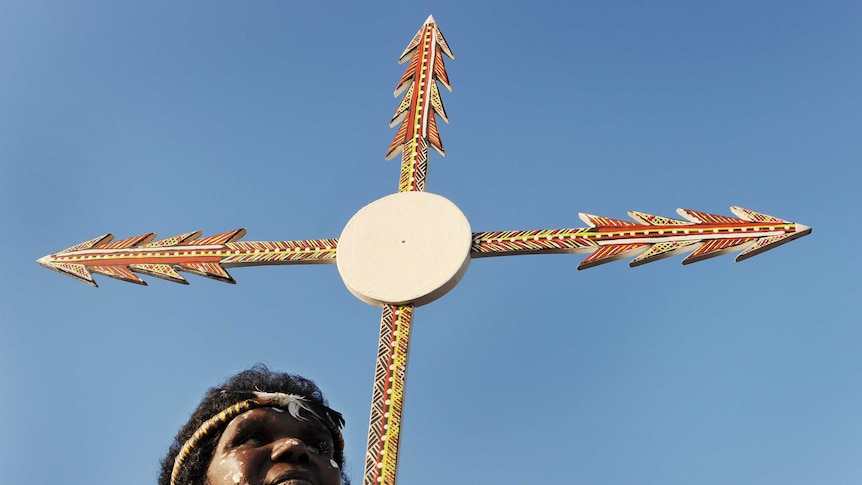 An Aboriginal woman holding a Christian cross made out of spears and painted in an Indigenous style.
