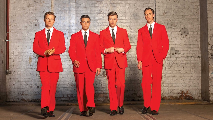Four men in red suits with black ties stand in front of a white brick wall.