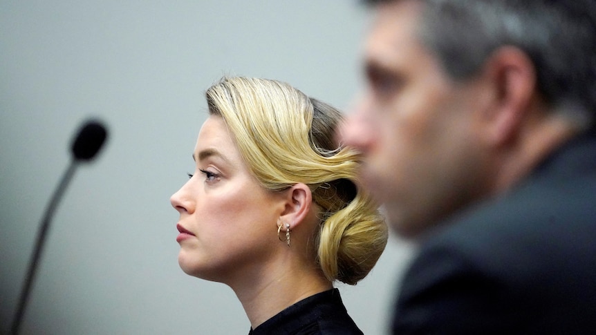 Side view of Amber Heard with hair pulled back, a mic on desk and a man blurred out