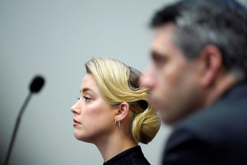 Side view of Amber Heard with hair pulled back, a mic on desk and a man blurred out