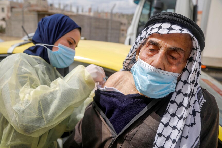 A Palestinian man in a mask and dishdash getting a vaccine from a health worker 