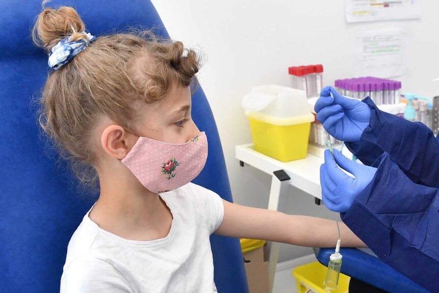 Blonde, curly-haired girl, about 8, wearing pink mask with nurse in blue smock, gloves, glasses getting medical test