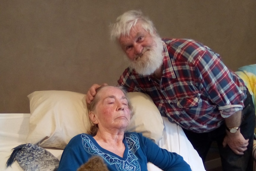 An elderly man and woman, the woman prone in bed.