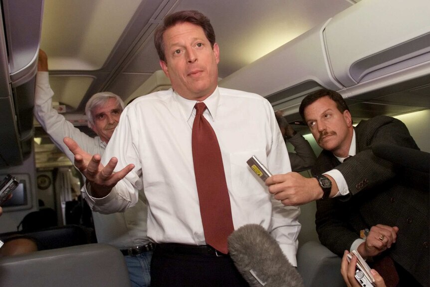 Gore shrugs as he's asked a question by a journalist leaning over a plane seat