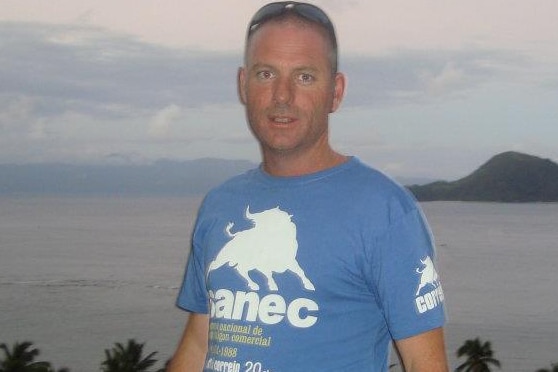 A mid shot of Nathan Haynes posing for a photo in a blue shirt in front of the ocean.