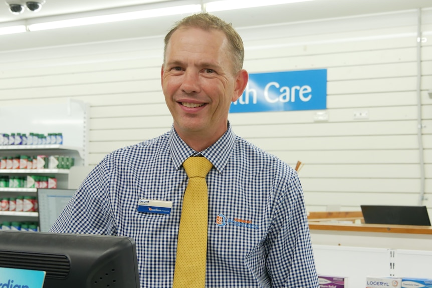 A man wearing a shirt with a logo, name badge, yellow tie, smiles as he stands behind the counter of a pharmacy.