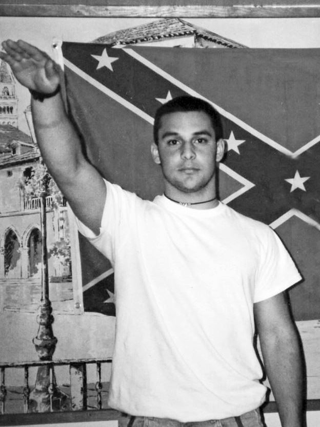 A man in white shirt and shaved head raises arm in Nazi salute, with neutral expression. Confederate flag is behind him.