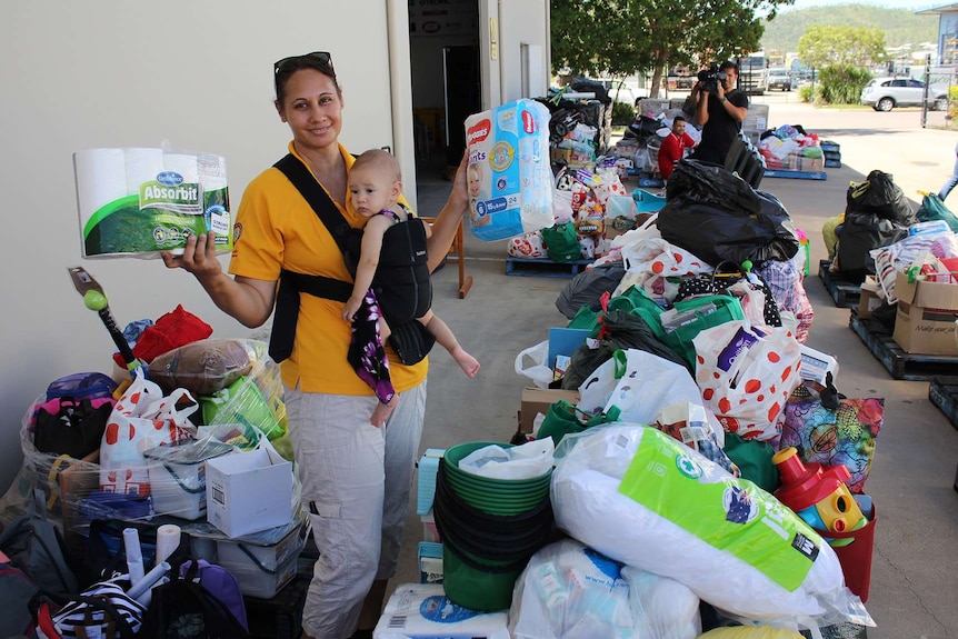 A woman stands holding nappies and toilet paper amongst a sea of donated goods for flood victims.