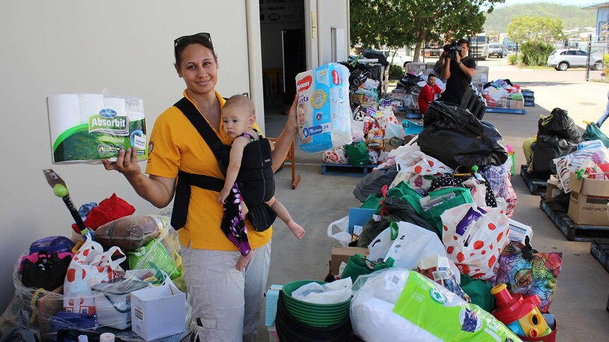 A woman stands holding nappies and toilet paper amongst a sea of donated goods for flood victims.