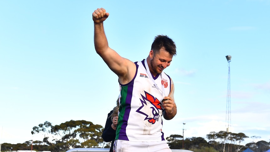 A man wearing a white football jumper holds a fist in the air with a medal around his neck.