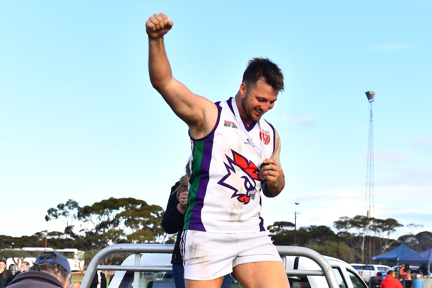 A man wearing a white football jumper holds a fist in the air with a medal around his neck.