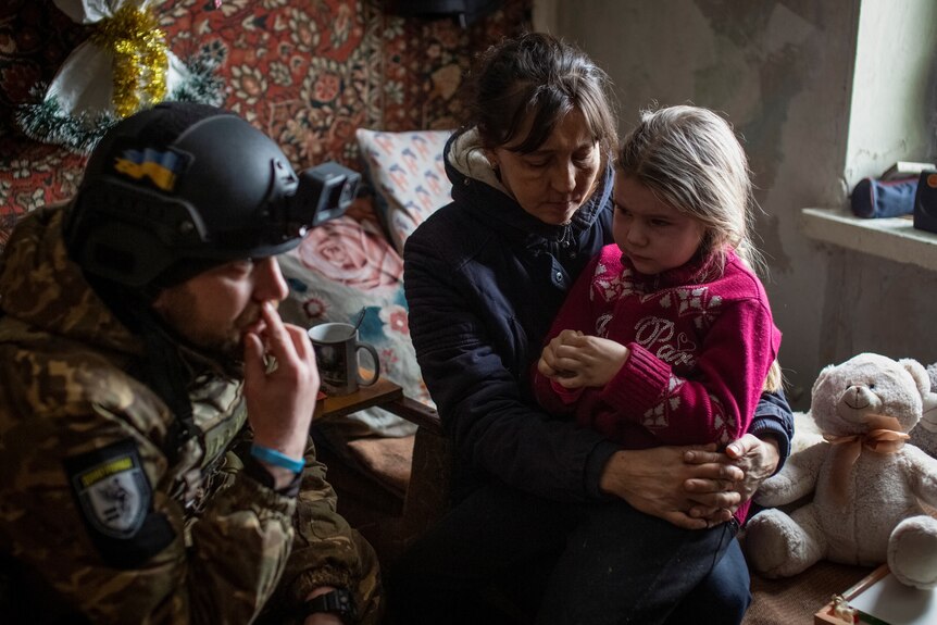 A girl sits on a woman's lap as a man in an army uniform speaks to her. 