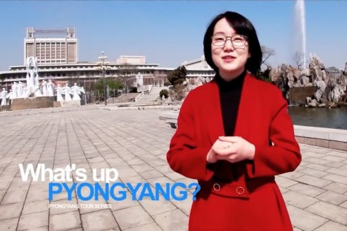 A young North Korean woman with glasses and a red jacket has her hands together in front of the camera.