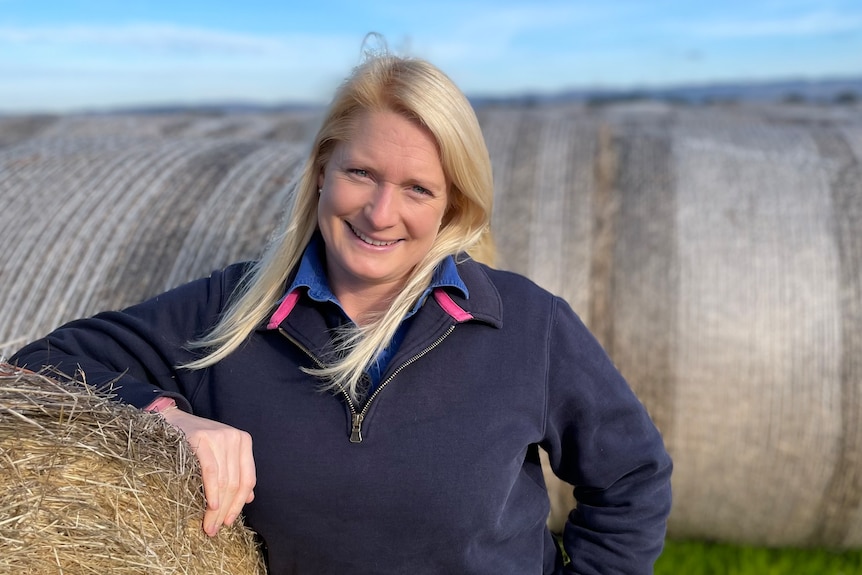 Woman with blonde hair and hand on a hay bale smiles at camera 