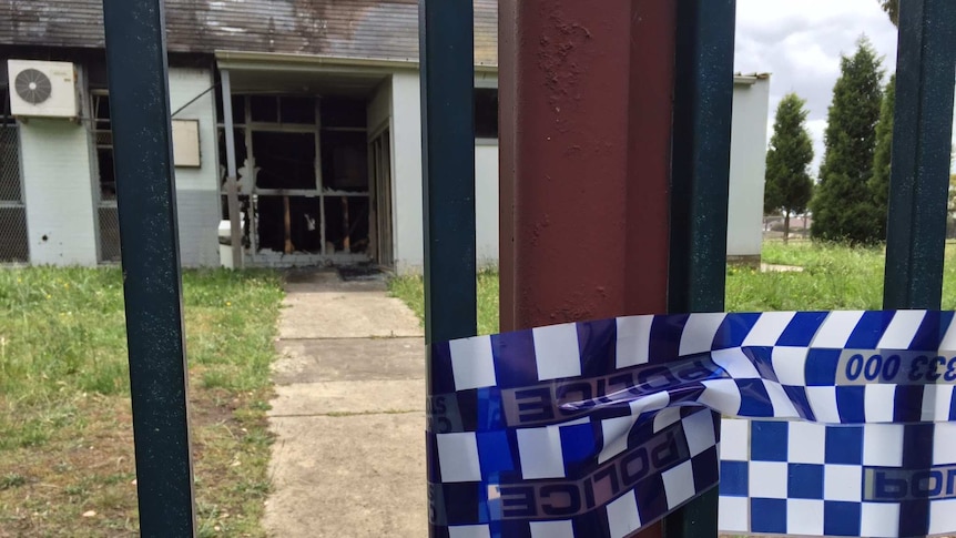 Police tape wrapped around a gate, with a burnt-out building in the background.
