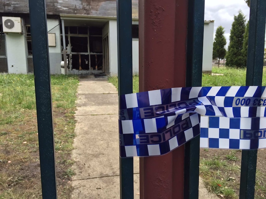 Police tape wrapped around a gate, with a burnt-out building in the background.