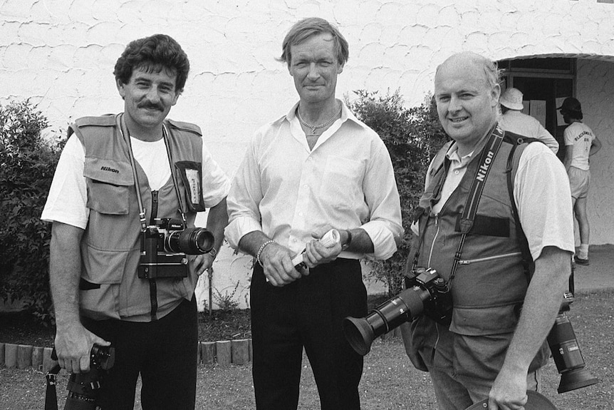 A black and white photograph of three men, two with cameras around their necks