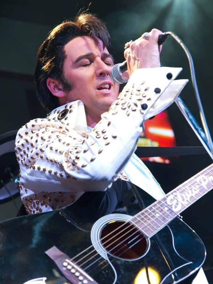 A man dressed as Elvis Presley sings into a microphone with his eyes closed