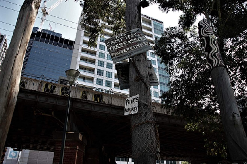 A picture of a rail bridge, with signs in front saying no camping and city highrise buildings behind.