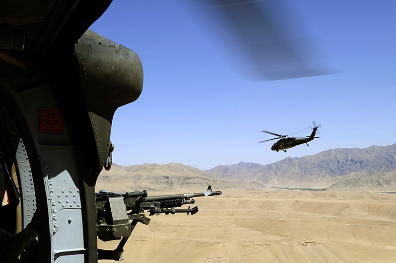 View of out of a helicopter, with gun in foreground, second helicopter and mountain range in distance