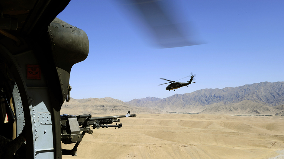 View of out of a helicopter, with gun in foreground, second helicopter and mountain range in distance