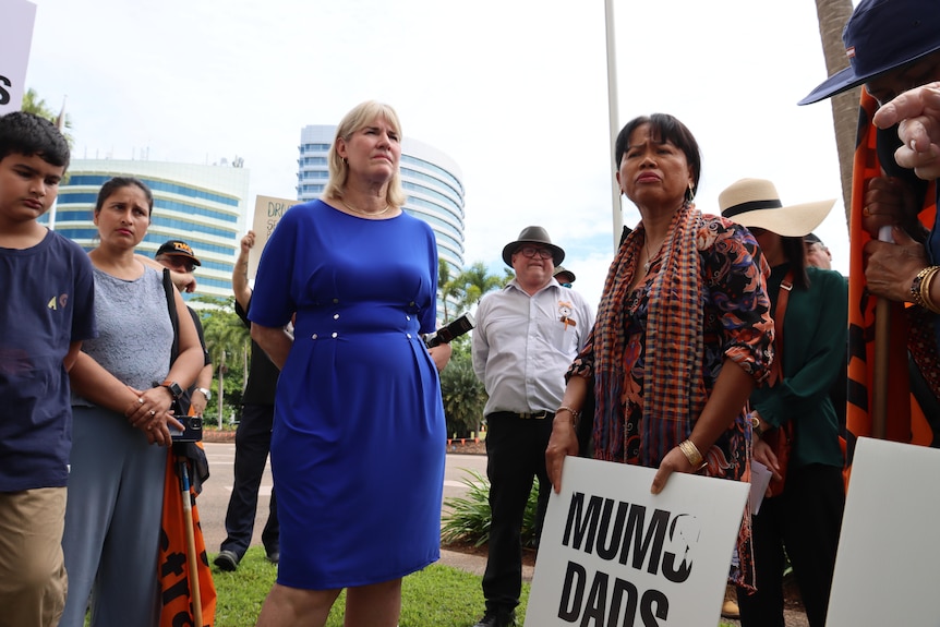 Minister Eva lawler stands looking solemn among a group of people, some holding signs. 