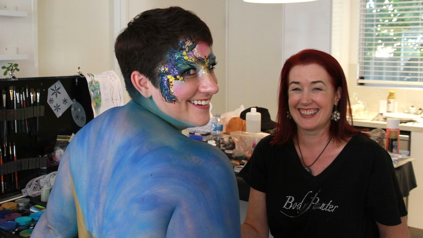 Covered in body paint Ria Cadell looks at camera with artist Wendy Fantasia in background.
